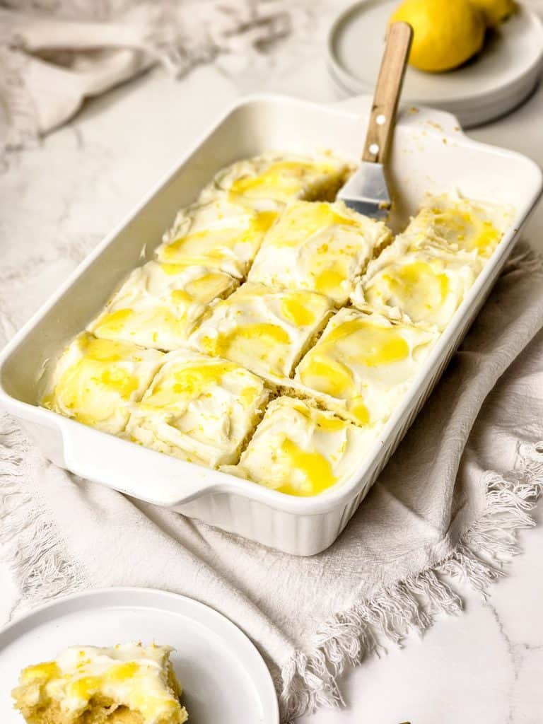 Rectangular baking dish with nine frosted lemon bars, some topped with lemon curd. A spatula rests on top. Nearby are empty plates and a whole lemon on a light textured surface.