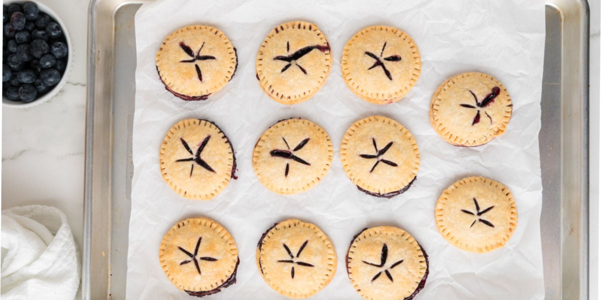 A baking tray lined with parchment paper holds twelve round mini pies with slotted tops; a bowl of blueberries is placed nearby. A baking tray lined with parchment paper holds twelve round mini pies with slotted tops; a bowl of blueberries is placed nearby.