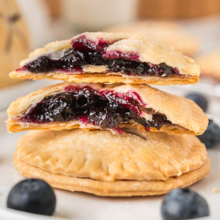 A stack of hand pies filled with blueberry filling, with the top pie split open to reveal the jammy interior; fresh blueberries are scattered nearby.