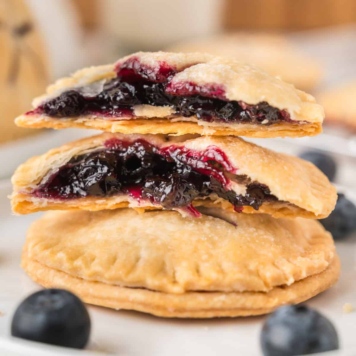 A stack of hand pies filled with blueberry filling, with the top pie split open to reveal the jammy interior; fresh blueberries are scattered nearby.