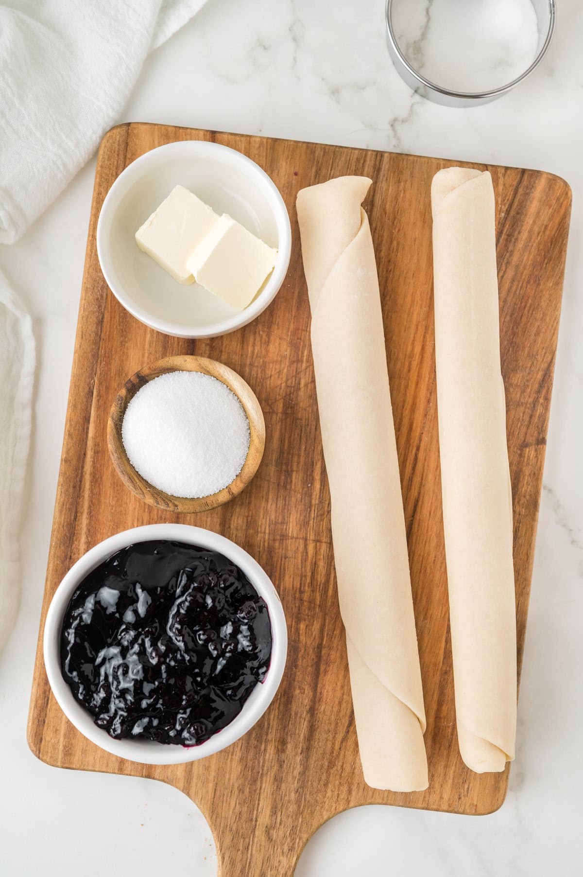 A wooden cutting board with rolled pie crusts, a bowl of butter, a bowl of sugar, and a bowl of blueberry preserves on a marble countertop.
