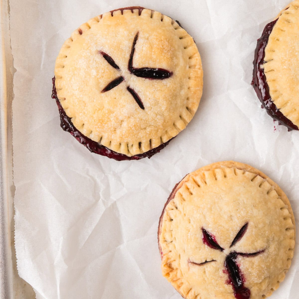 Three round blueberry pie cookies with crimped edges and blueberry filling are on a parchment-lined baking sheet. Two are fully visible, and one is partially shown.