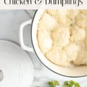 White Dutch oven filled with chicken and dumplings on a marble countertop, garnished with fresh parsley; lid placed to the side.
