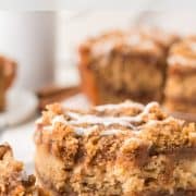 A slice of cinnamon coffee cake with crumb topping and icing, served on a white plate with more slices in the background.