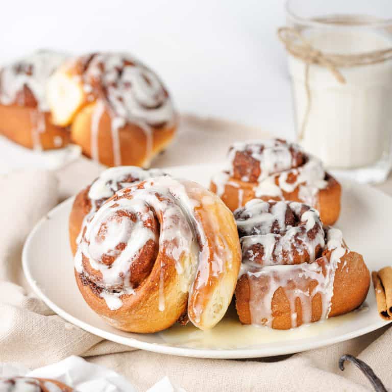 A plate of cinnamon rolls drizzled with icing, with more rolls and a glass of milk in the background.