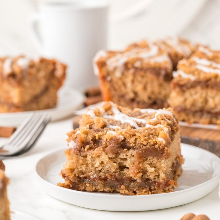 A square piece of cinnamon coffee cake with icing over crumble topping on a white plate, with more cake pieces and a white mug in the background.