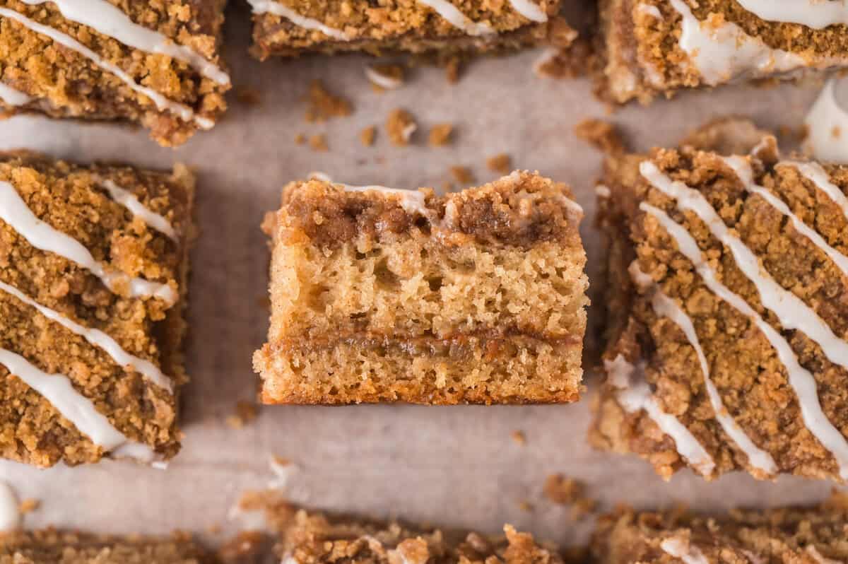 A rectangular piece of coffee cake with a crumb topping and icing, surrounded by similar pieces on a parchment paper surface. A rectangular piece of coffee cake with a crumb topping and icing, surrounded by similar pieces on a parchment paper surface.