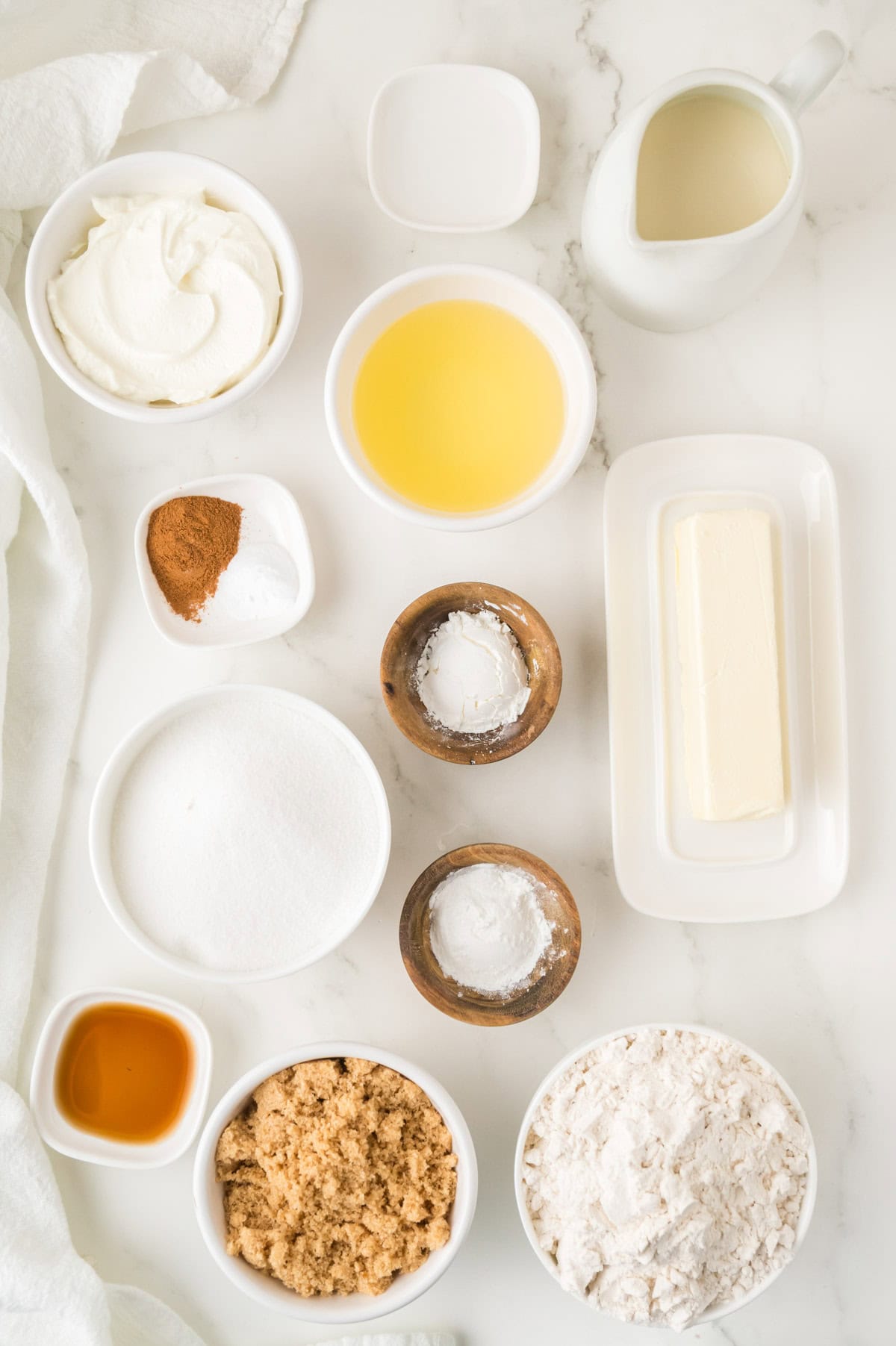 Various baking ingredients, including flour, sugar, brown sugar, butter, vanilla extract, cream cheese, milk, cinnamon, and wet ingredients, arranged on a white marble surface.
