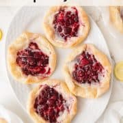 Four raspberry danishes with icing are arranged on a white plate, with a bowl of raspberry jam and part of a lemon visible in the background on a marble surface.