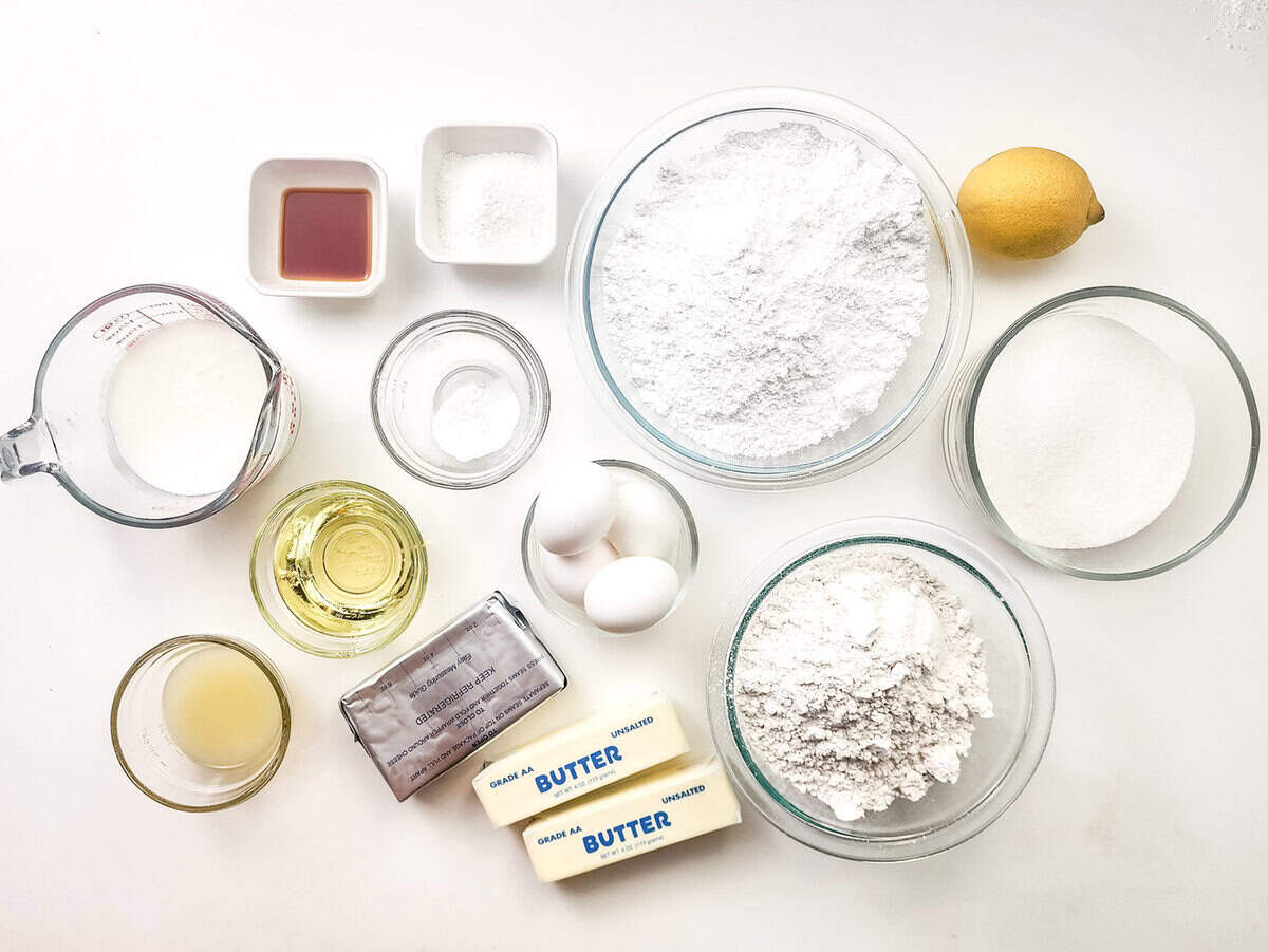 Various baking ingredients, including flour, sugar, eggs, butter, oil, lemon, vanilla extract, milk, and baking powder, arranged in bowls and cups on a white surface. Various baking ingredients, including flour, sugar, eggs, butter, oil, lemon, vanilla extract, milk, and baking powder, arranged in bowls and cups on a white surface.