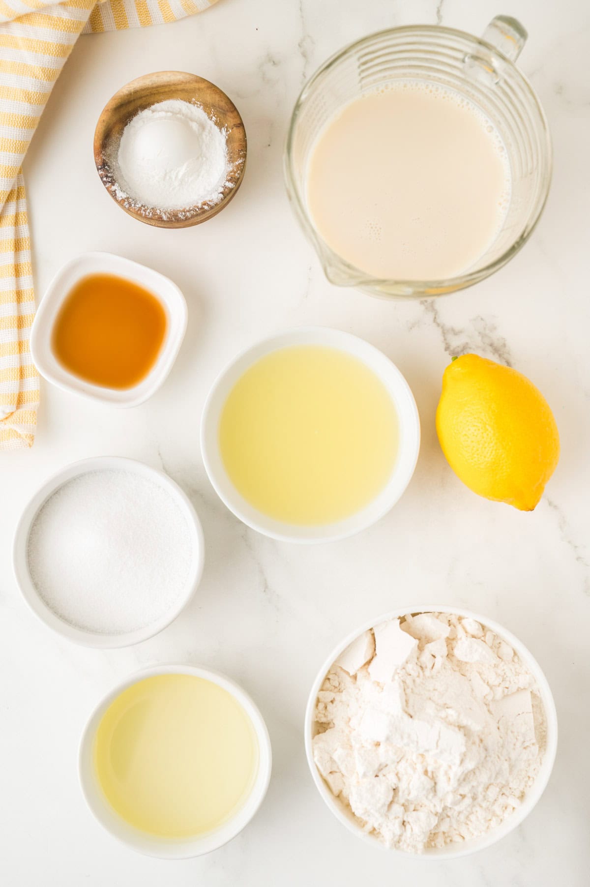 Ingredients for pancakes on a white surface including flour, sugar, oil, lemon, vanilla, baking powder, and a measuring cup of milk.