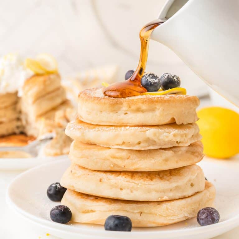 A stack of thick lemon pancakes topped with blueberries being drizzled with syrup, with more pancakes and a lemon in the background.