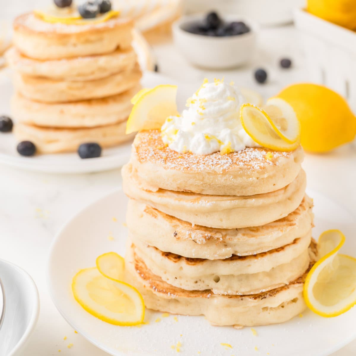 A stack of thick pancakes topped with whipped cream, lemon slices, and powdered sugar, with blueberries and a lemon in the background.