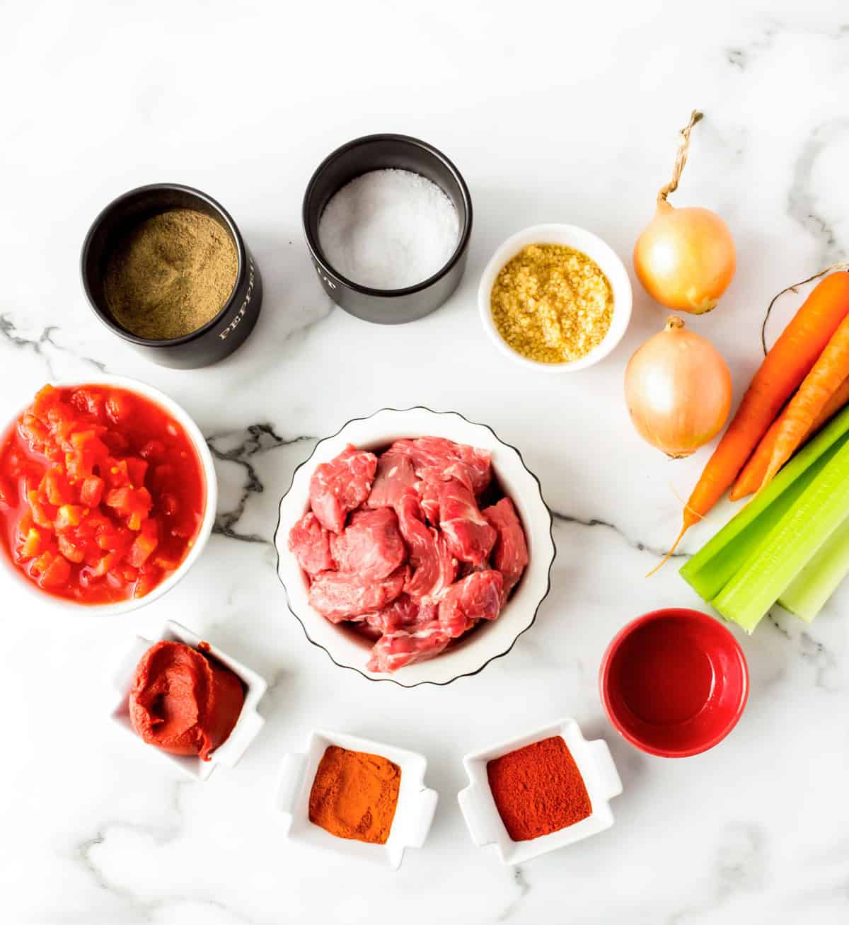 Raw beef cubes, chopped tomatoes, tomato paste, chopped garlic, spices, salt, pepper, onions, carrots, celery, and a small bowl of red liquid arranged on a white marble surface.