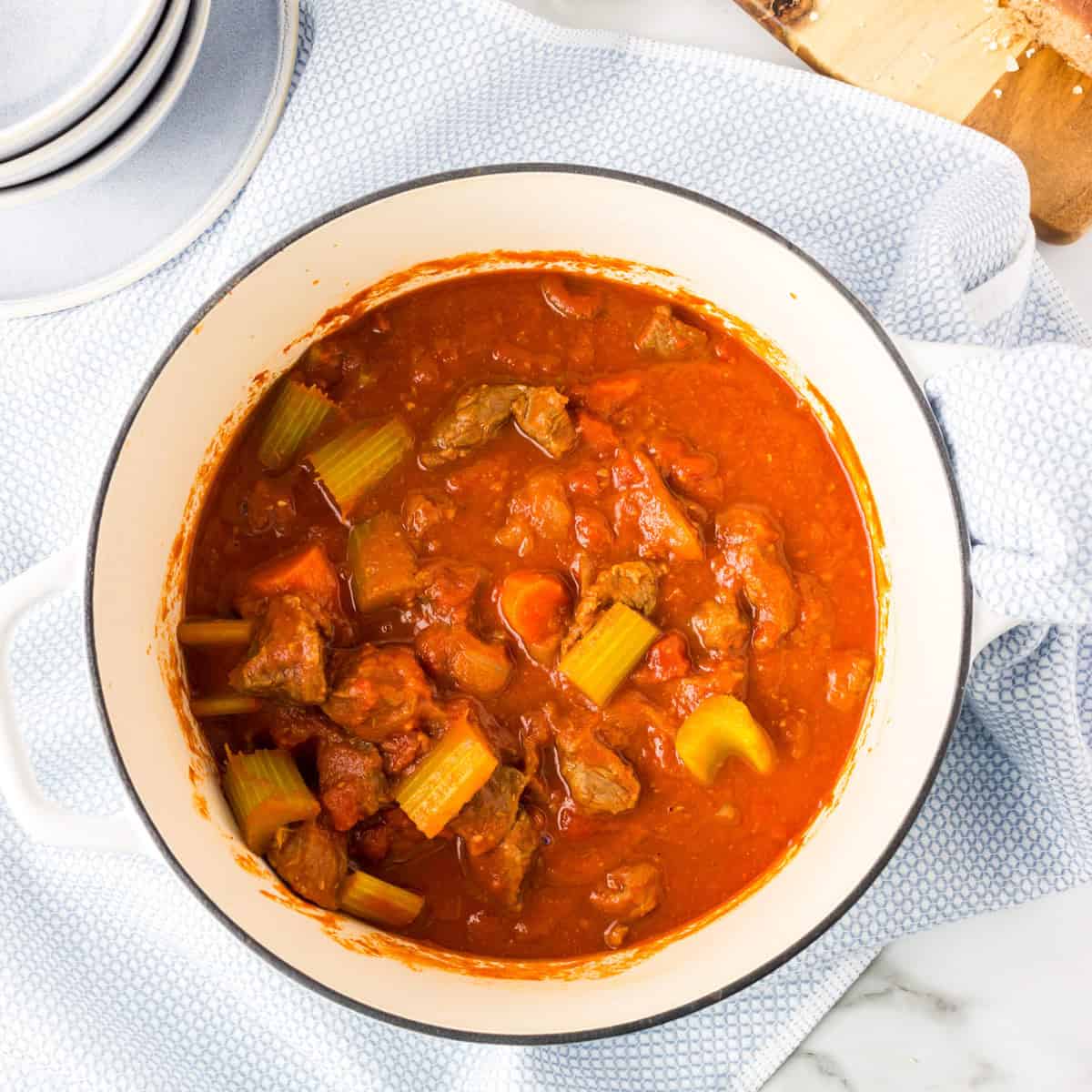 A pot of paprika beef stew with carrots and celery, surrounded by raw onions, celery stalk, and carrots on a white marble surface.