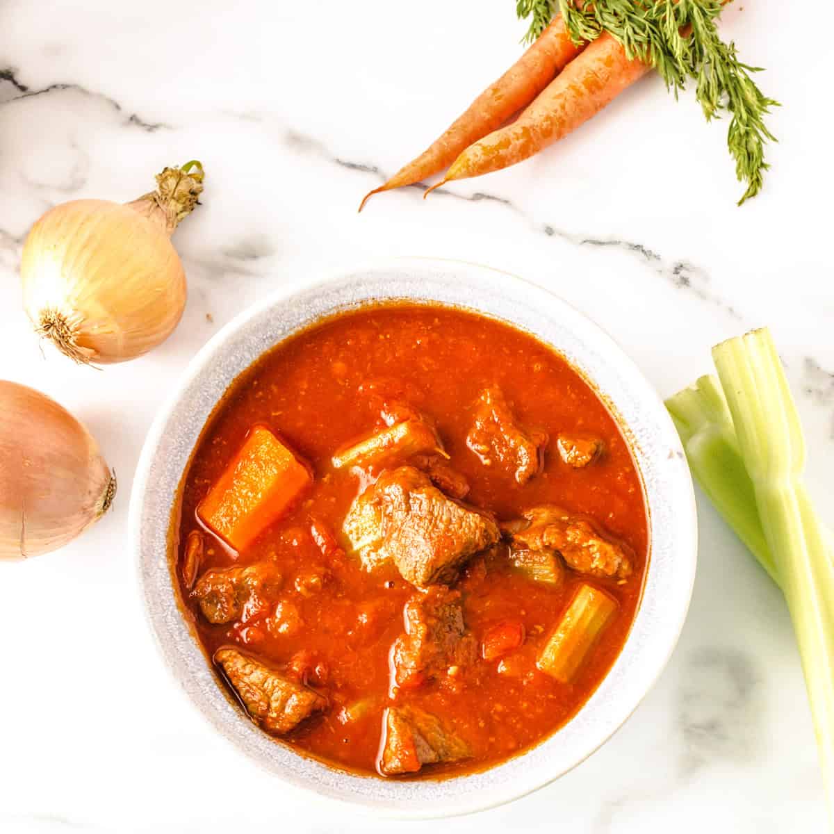 A bowl of beef stew with carrots and celery, surrounded by raw onions, celery stalk, and carrots on a white marble surface.
