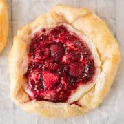 A puff pastry berry danish with flak, filled with raspberries and jam, sits on parchment paper. A rustic berry tart with flaky pastry crust, filled with mixed berries and jam, sits on parchment paper.