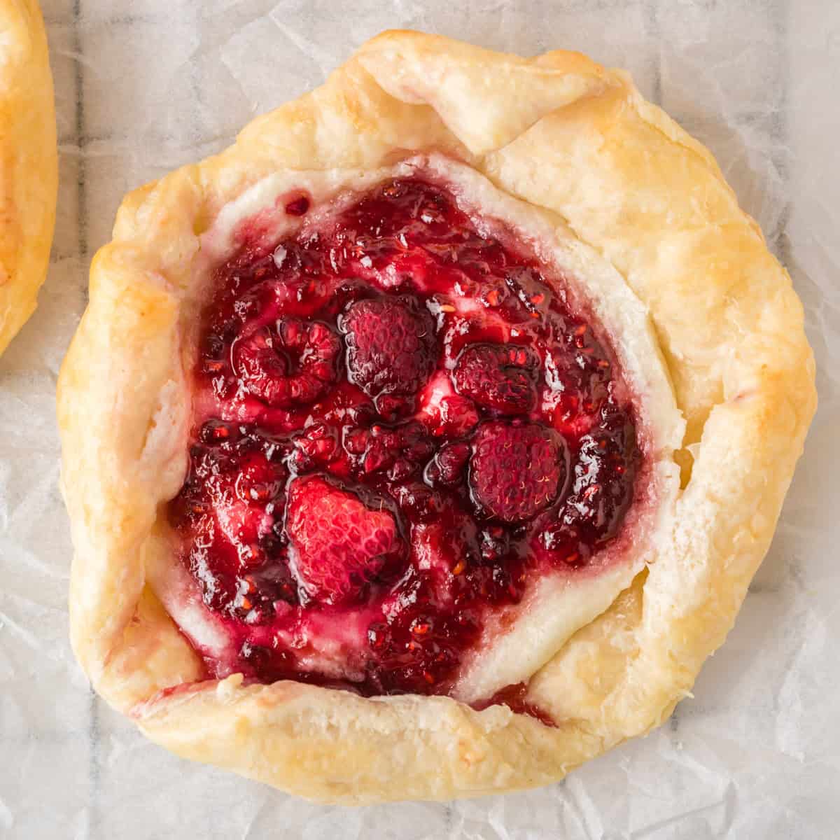 A puff pastry berry danish with flak, filled with raspberries and jam, sits on parchment paper. A rustic berry tart with flaky pastry crust, filled with mixed berries and jam, sits on parchment paper.