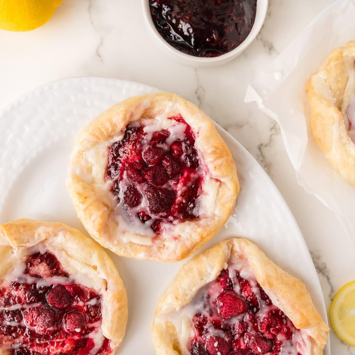 Three raspberry danishes with icing are arranged on a white plate, with a bowl of raspberry jam and part of a lemon visible in the background on a marble surface.