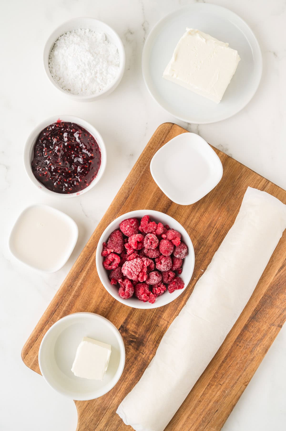 Ingredients on a countertop include cream cheese, powdered sugar, raspberry jam, frozen raspberries, butter, milk, and a rolled sheet of dough on a wooden cutting board.