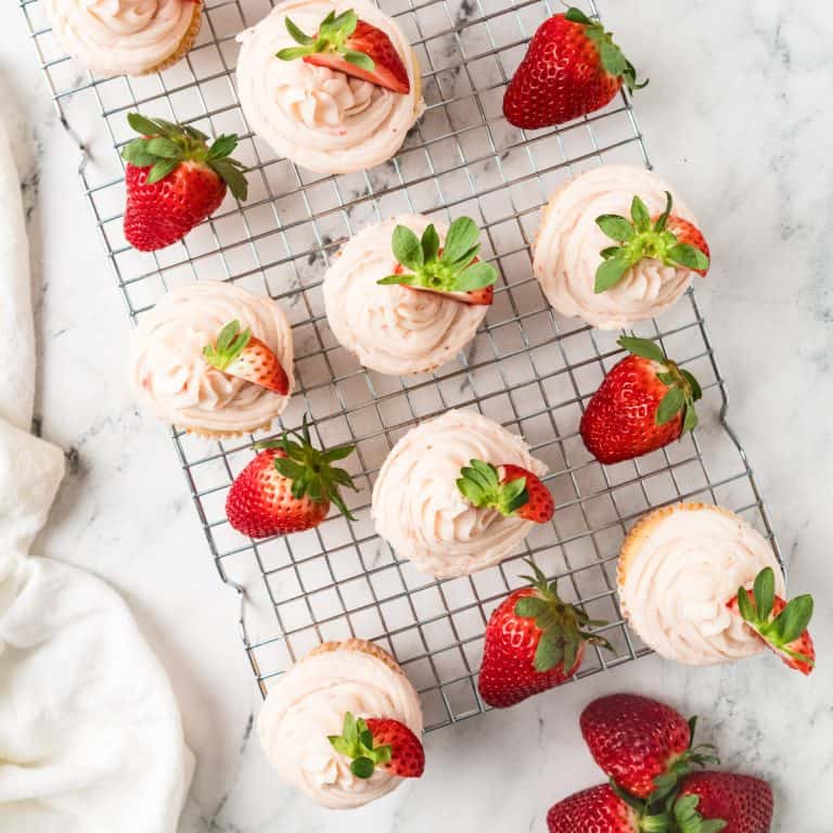 Strawberry cupcakes with frosting and fresh strawberries are arranged on a wire cooling rack on a marble surface. Cupcakes with pink frosting and fresh strawberries are arranged on a wire cooling rack on a marble surface.