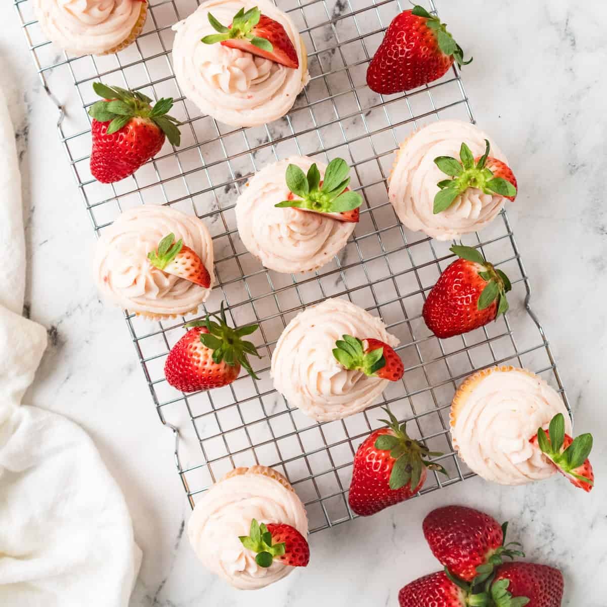 Strawberry cupcakes with frosting and fresh strawberries are arranged on a wire cooling rack on a marble surface. Cupcakes with pink frosting and fresh strawberries are arranged on a wire cooling rack on a marble surface.