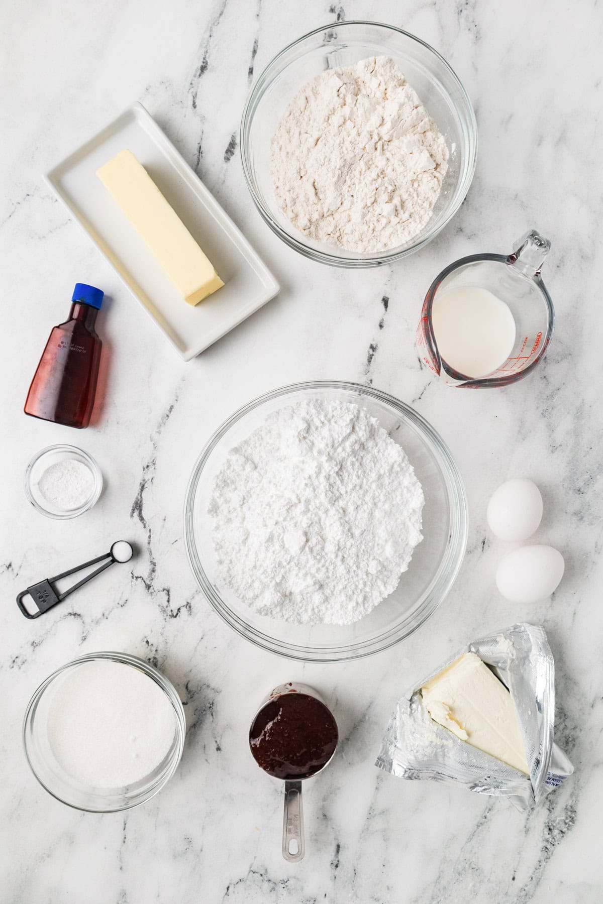 Various baking ingredients, including flour, sugar, powdered sugar, butter, cream cheese, eggs, vanilla extract, cocoa, baking soda, and milk, arranged on a marble surface.