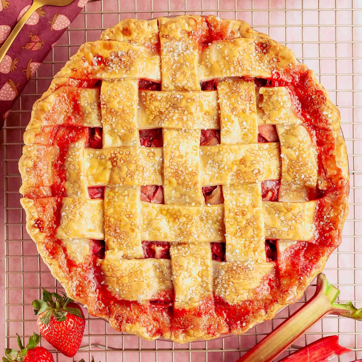 A lattice-topped strawberry rhubarb pie sits on a pink surface with fresh strawberries, and rhubarb.