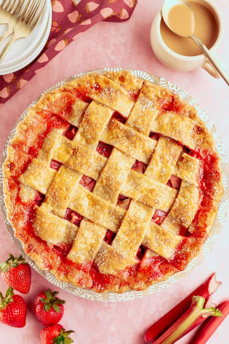 A lattice-topped strawberry rhubarb pie sits on a pink surface with fresh strawberries, rhubarb, a jug of sauce, and a stack of plates nearby.