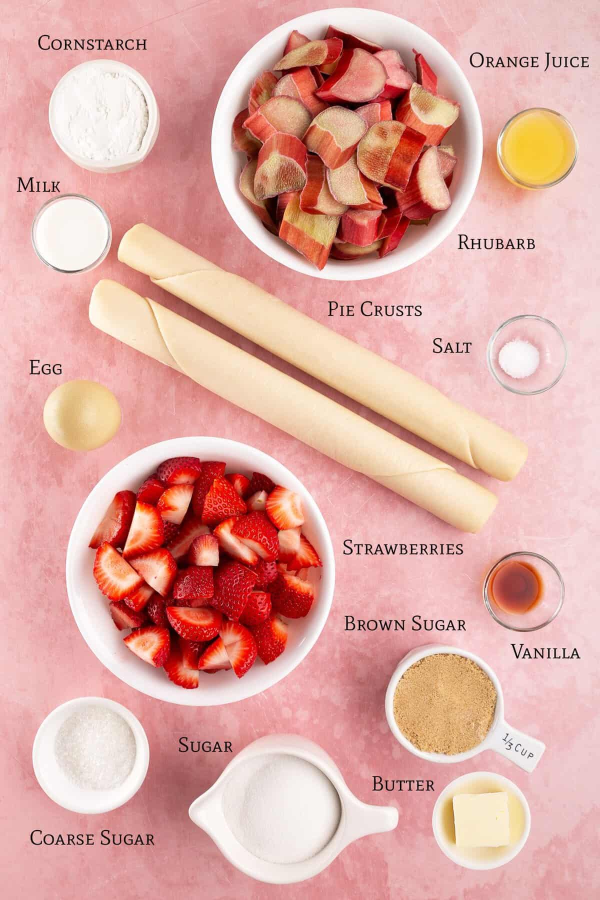 Ingredients for a strawberry rhubarb pie arranged on a pink surface, including rhubarb, strawberries, pie crusts, sugars, butter, egg, cornstarch, vanilla, milk, orange juice, and salt.