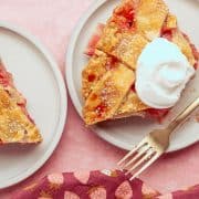 Two slices of strawberry rhubarb pie on plates, one topped with whipped cream, beside a gold fork and a strawberry-patterned cloth on a pink surface.