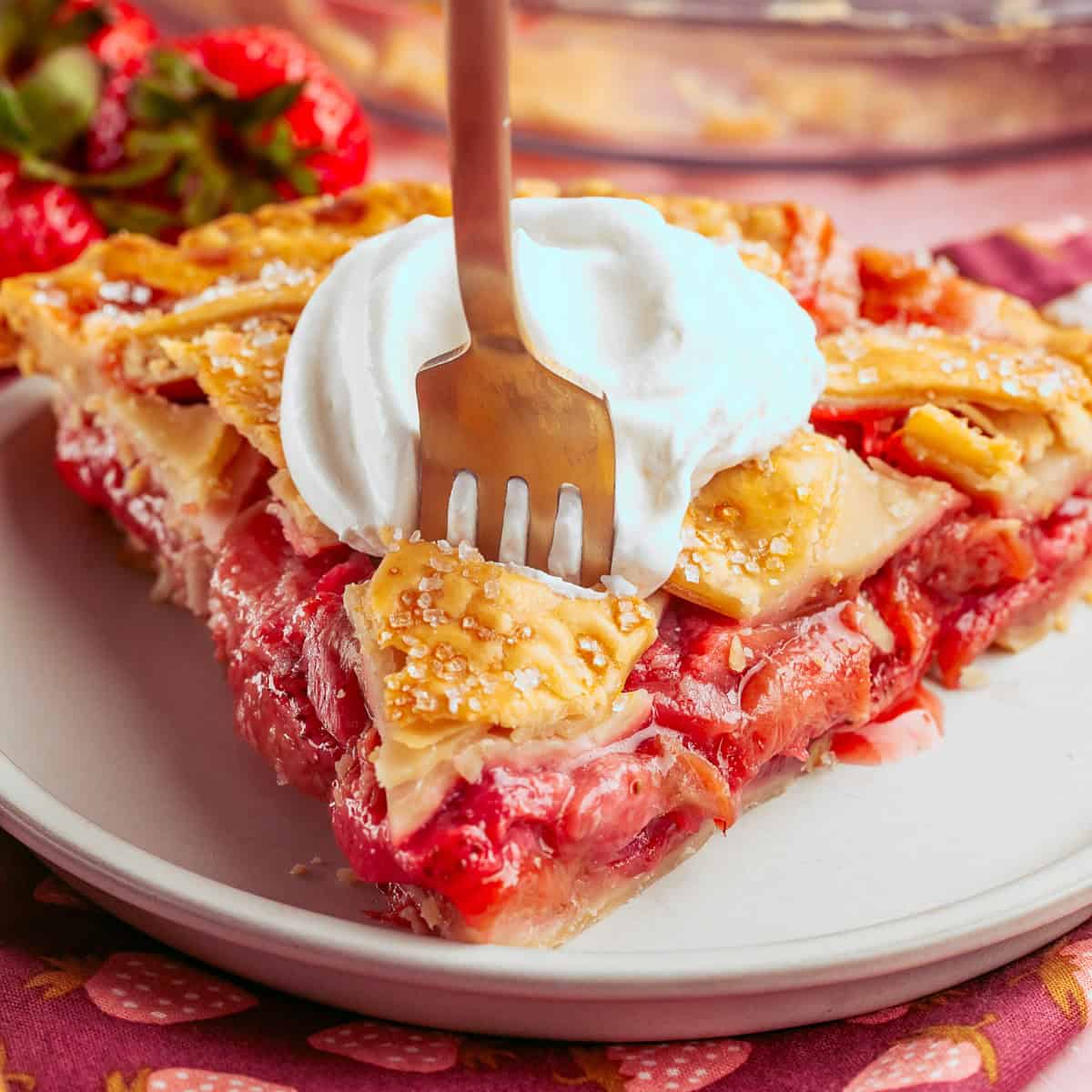 A fork cutting into a slice of strawberry rhubarb pie topped with whipped cream on a white plate.