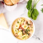 A bowl of creamy tortellini soup with sausage, spinach, and tomatoes, surrounded by bread rolls, fresh spinach, a cheese wedge, and herbs on a marble surface.