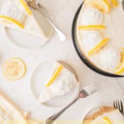 Top-down view of a no-bake lemon cheesecake garnished with lemon slices and whipped cream, with two plated slices, forks, and lemon halves on a light background. Text reads No-Bake Lemon Cheesecake.