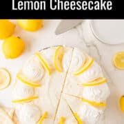 A no-bake lemon cheesecake topped with whipped cream and lemon slices, partially sliced, surrounded by whole and sliced lemons on a white surface.