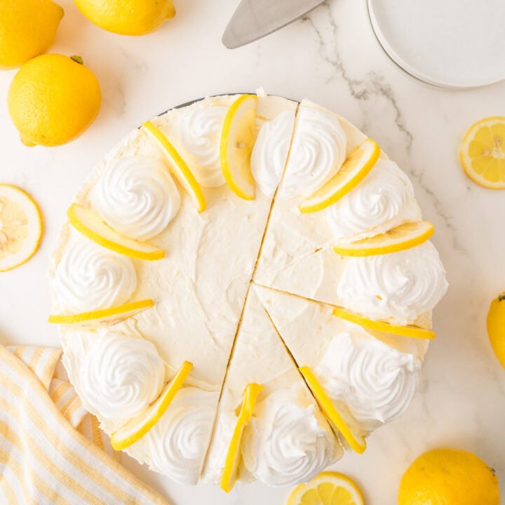 Top view of a no-bake lemon cheesecake topped with whipped cream swirls and lemon slices, partially sliced, surrounded by whole and sliced lemons on a marble surface.