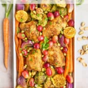Roasted chicken thighs with carrots, Brussels sprouts, red onions, grapes, lemon slices, and garnished with chopped herbs and pistachios in a white rectangular dish.
