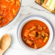 A bowl of paprika beef stew with vegetables, next to a pot of stew, a wooden spoon, and a cutting board with fresh carrots and celery.