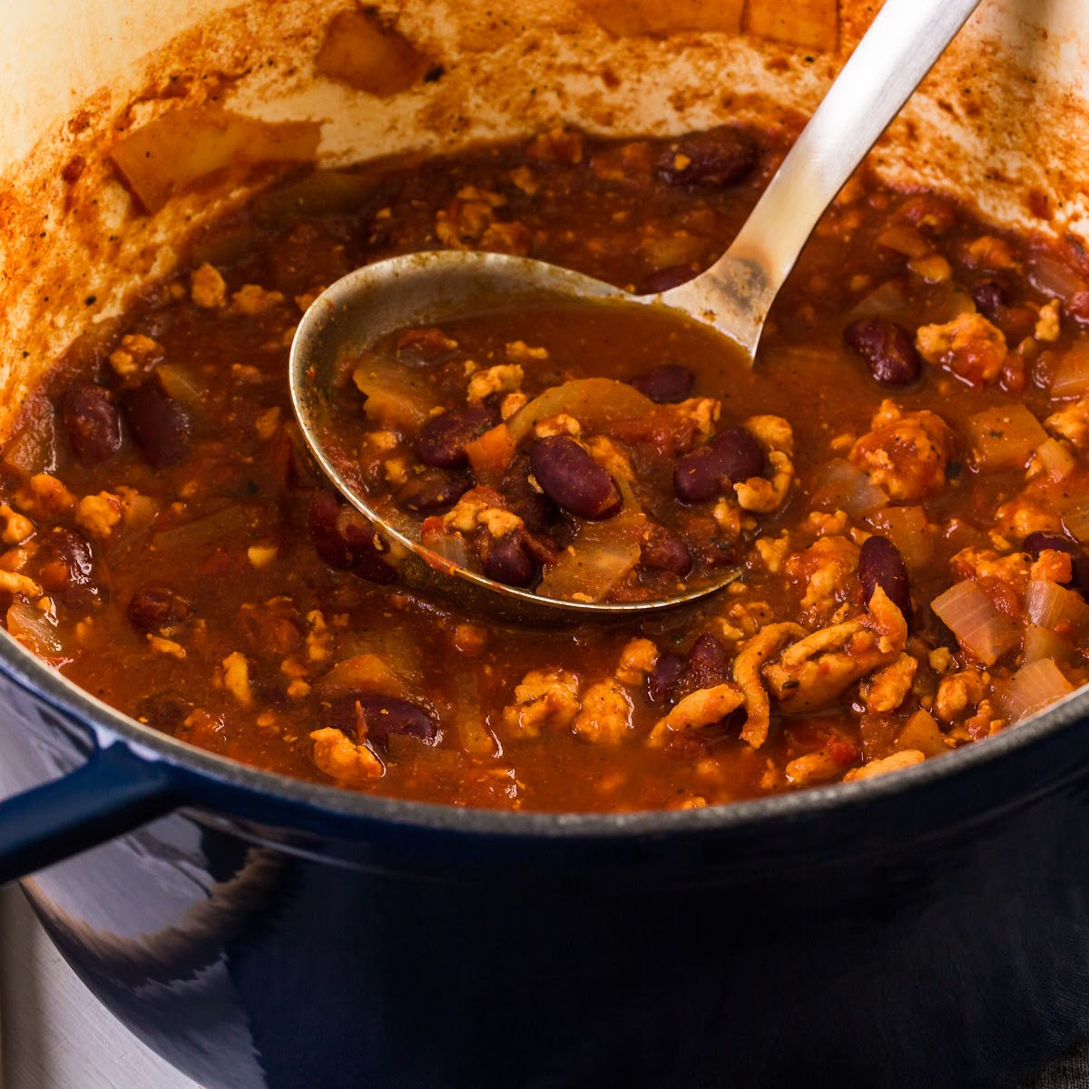 A ladle scoops chili with beans, ground meat, and onions from a blue pot.
