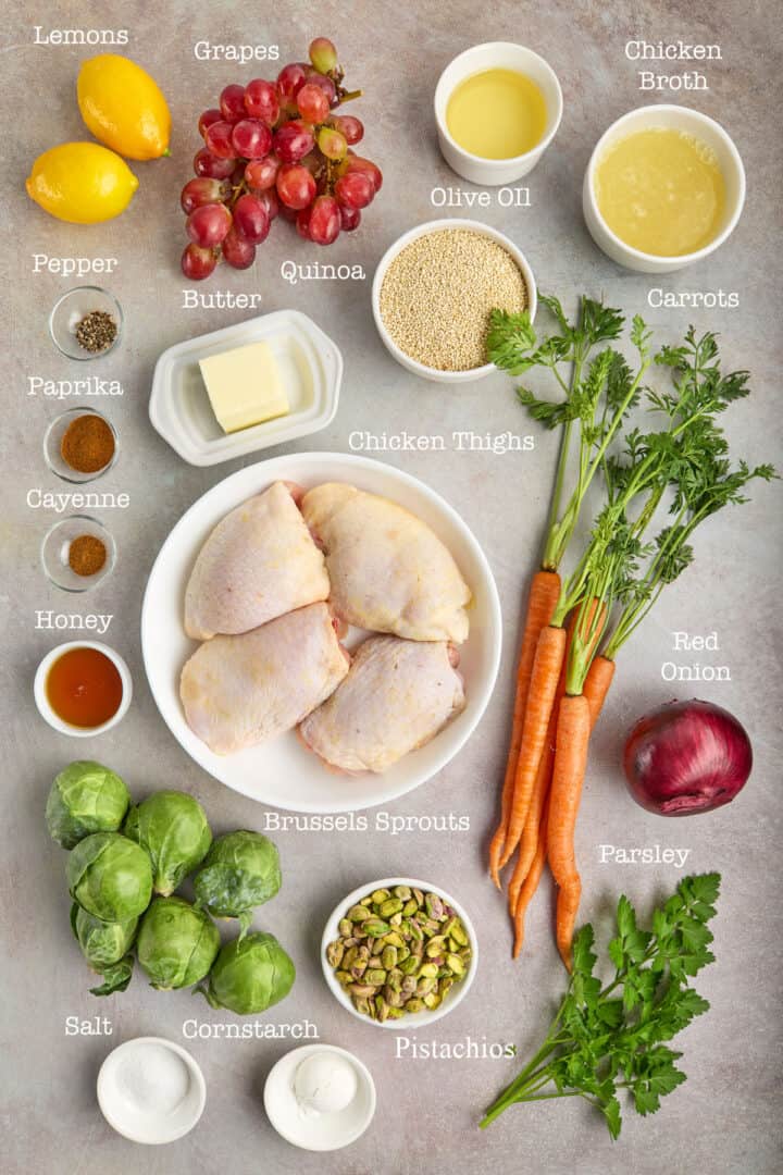 Assorted ingredients for a chicken dish, including chicken thighs, vegetables, spices, quinoa, olive oil, chicken broth, butter, honey, pistachios, lemon, and parsley, arranged on a table.