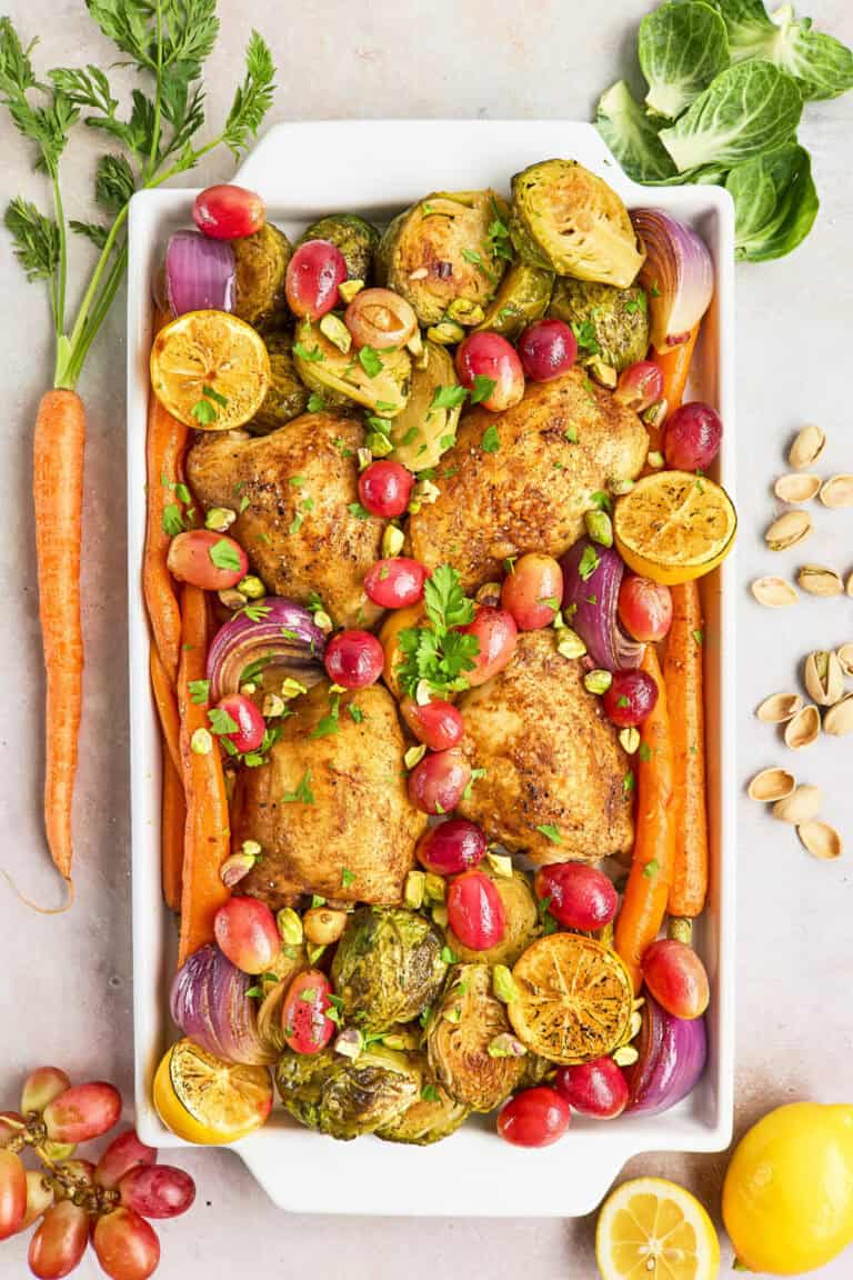 Baked chicken thighs, roasted carrots, Brussels sprouts, red onions, grapes, lemon slices, and garnished with chopped pistachios and parsley in a white baking dish.