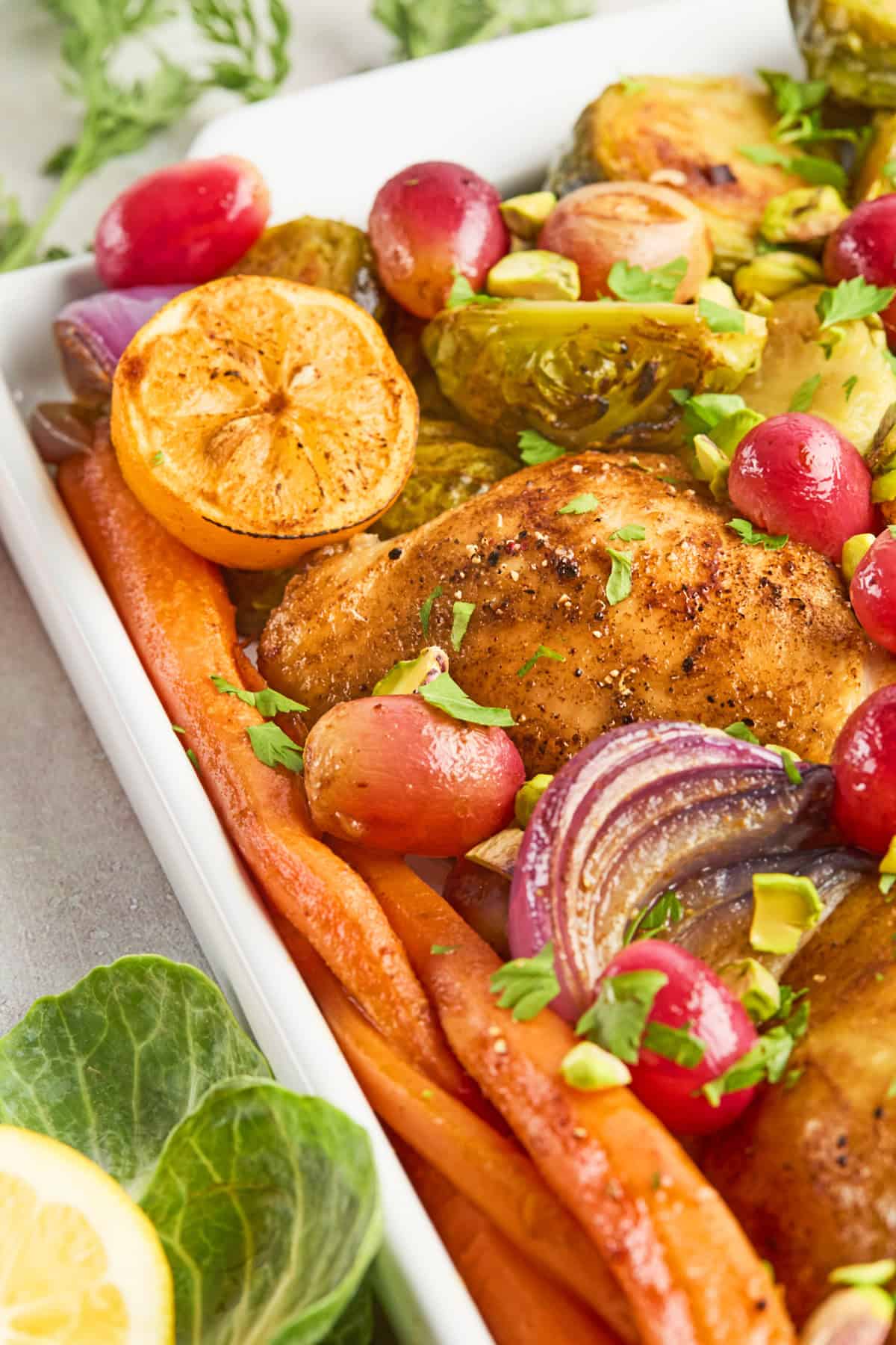 A roasted chicken breast with Brussels sprouts, carrots, red onions, halved lemons, grapes, and chopped herbs arranged in a white dish.