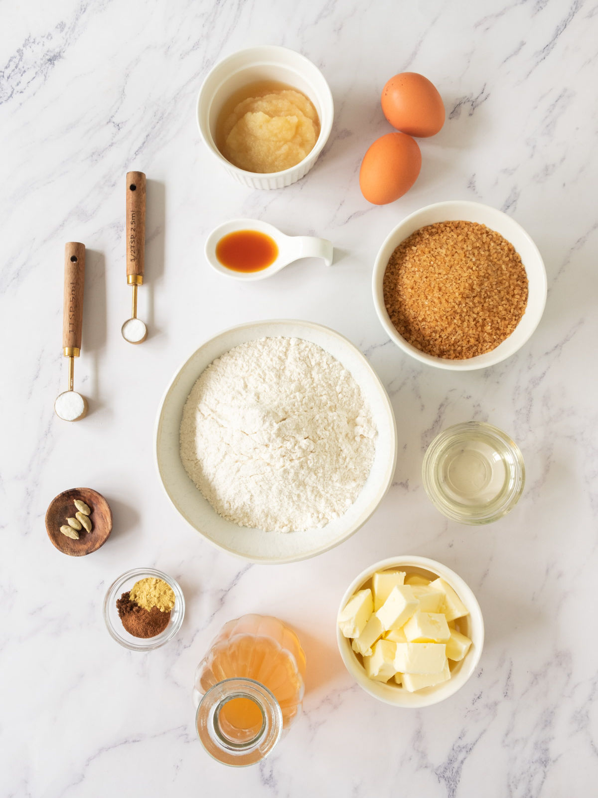 A flat lay of baking ingredients for apple cider bundt cake on a white marble surface, including flour, brown sugar, butter cubes, eggs, applesauce, spices, vanilla, oil, and measuring spoons with salt and baking powder.