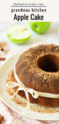 A bundt apple cake with a dusting of cinnamon sugar on top, tied with a white ribbon, sits on a plate. A green apple, some apple slices, and cinnamon sticks are in the background.