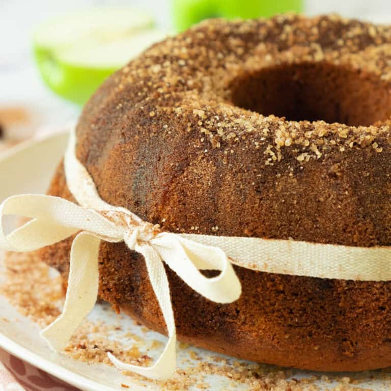 A bundt cake topped with cinnamon sugar sits on a plate. The cake is wrapped with a cream-colored ribbon tied in a bow. Crumbs and pieces of sugar are scattered on the plate.