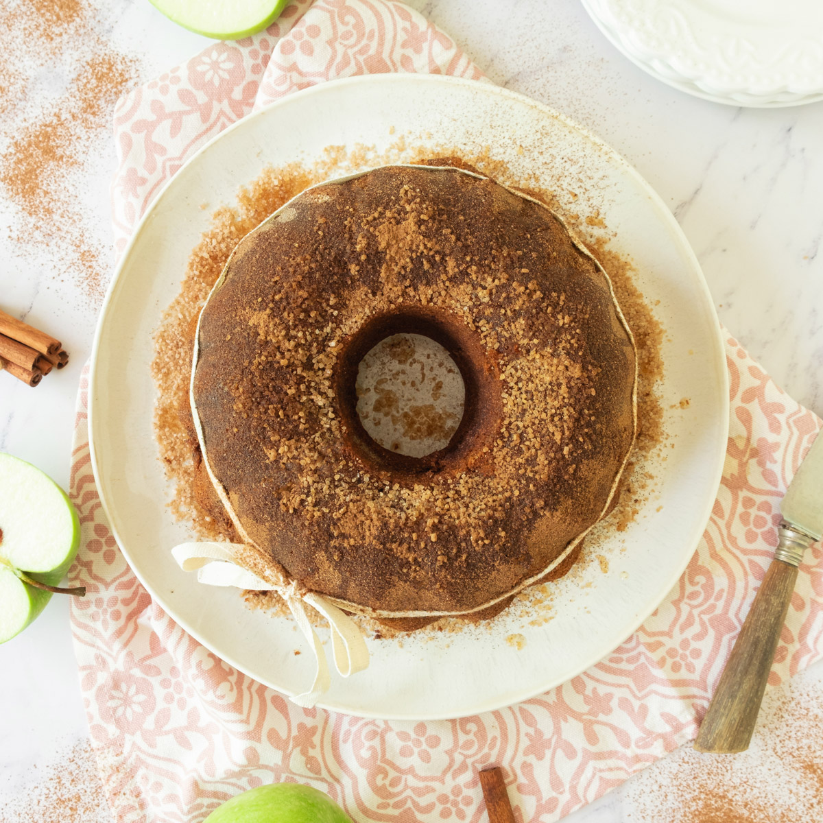 A round Bundt cake dusted with cinnamon and brown sugar sits on a white plate atop a patterned cloth, surrounded by apple slices, cinnamon sticks, and a cake server on a marble surface.