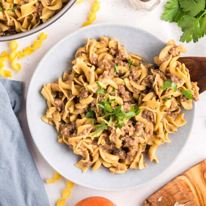 A plate of creamy beef stroganoff with egg noodles, garnished with parsley, sits on a white surface with a wooden spoon and uncooked pasta nearby.