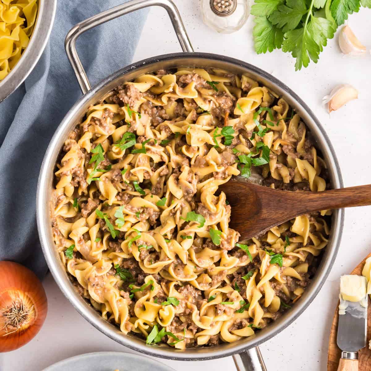 Large skillet filled with creamy beef stroganoff pasta garnished with fresh parsley. A wooden spoon is in the skillet, surrounded by ingredients like an onion, parsley, garlic, and uncooked egg noodles on the table.