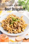 A plate of creamy beef stroganoff with egg noodles, garnished with parsley, sits on a table. Surrounding items include a pot, an onion, parsley, and a knife. Text reads Classic & Creamy Beef Stroganoff.