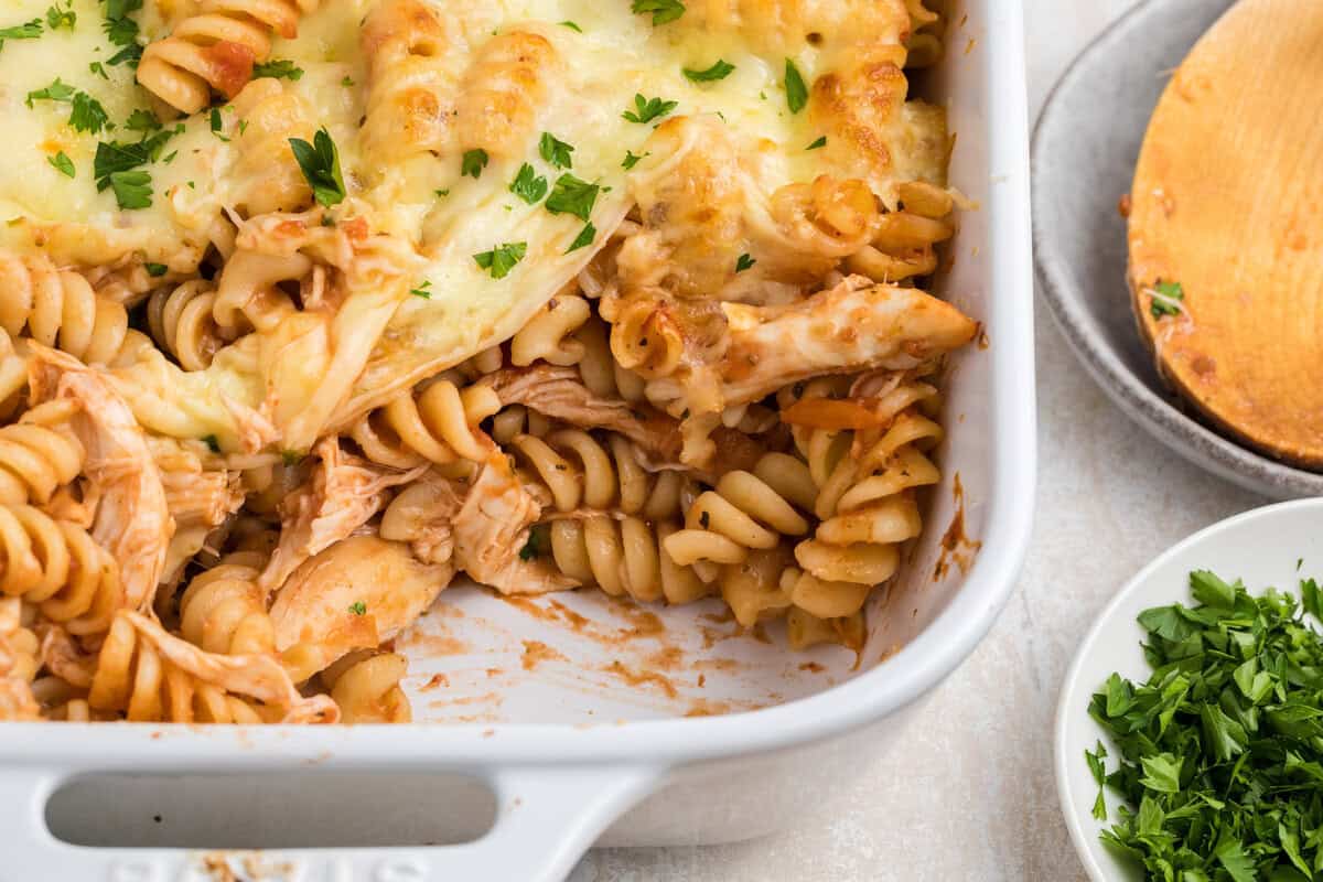 Baked rotini pasta with shredded chicken and melted cheese, garnished with chopped parsley, in a white casserole dish.